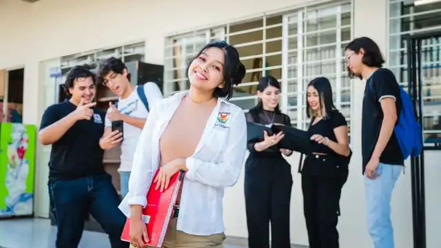 Estudantes em campus universitário; jovem no primeiro plano sorri e segura um caderno vermelho